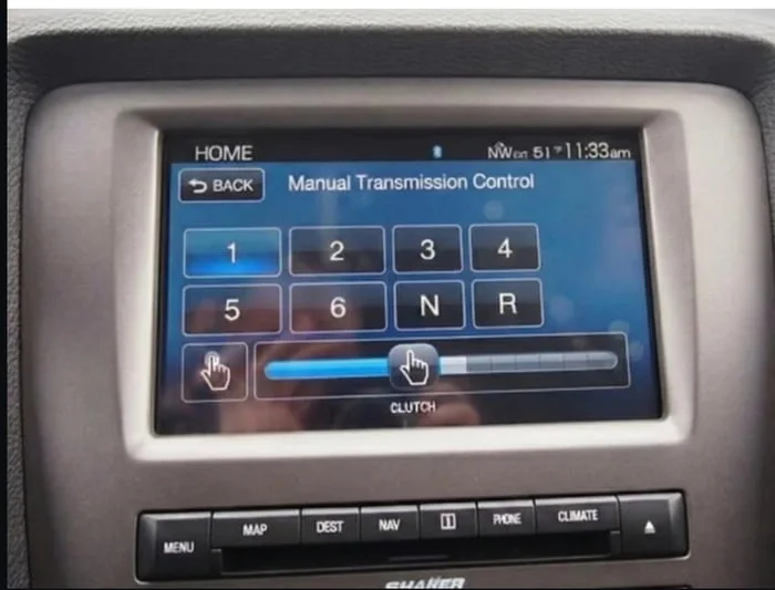 How to drive manual in 2030. In-car touchscreen display showing a manual transmission control interface with gear selection buttons. Text: HOME / BACK / Manual Transmission Control / 1 2 3 4 / 5 6 N R / CLUTCH / MENU MAP DEST NAV MODE CLIMATE / SHAKER
