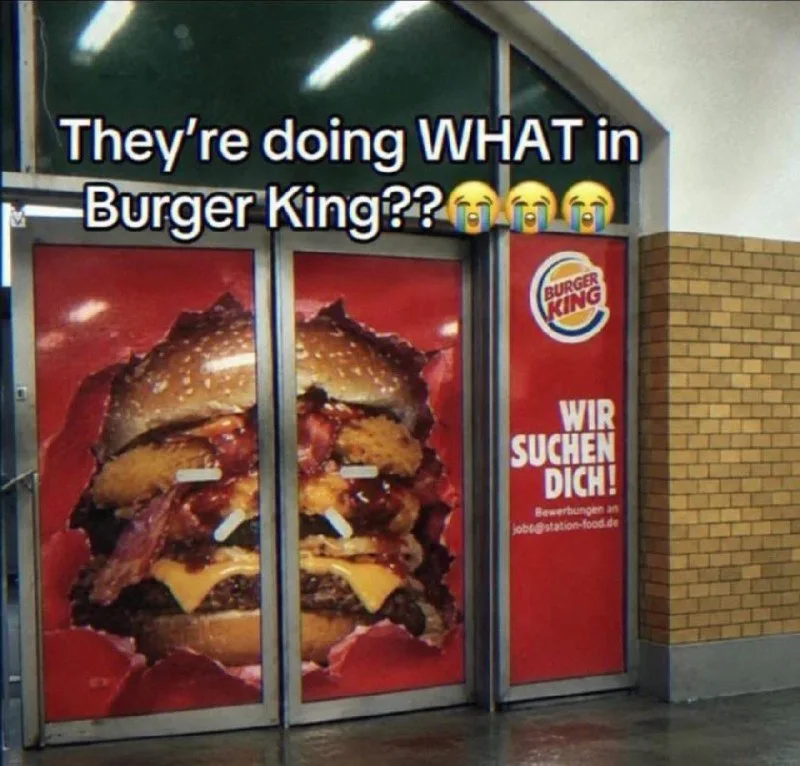 Burger King advertisement window showing a large burger with the German text 'Wir suchen dich' (We're hiring you). Text: They're doing WHAT in Burger King?? / BURGER KING / WIR SUCHEN DICH! / Bewerbungen an jobs@station-food.de