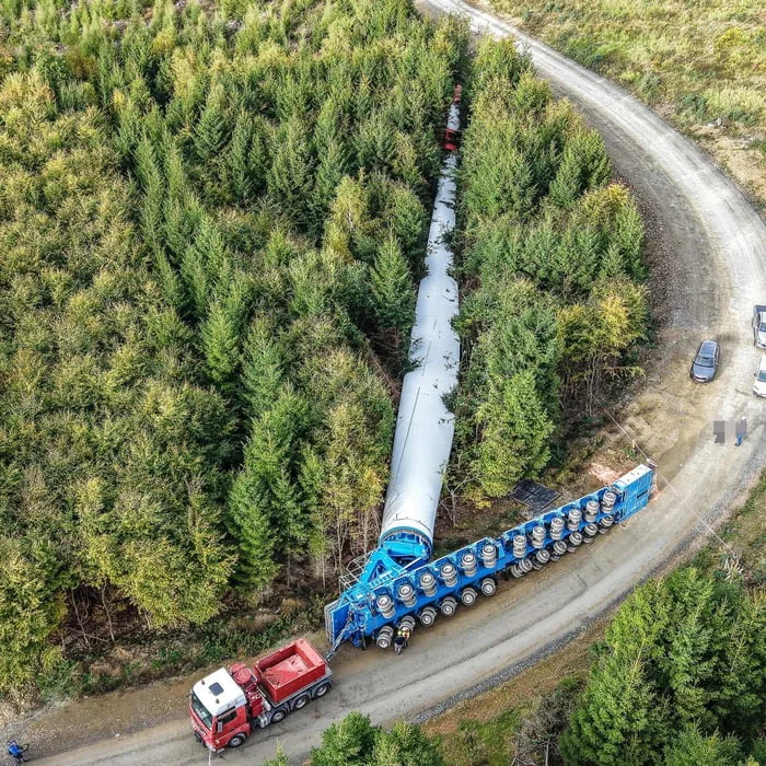 Looks like someone missed their turn. Aerial view of an oversized wind turbine blade being transported on a specialized trailer through a forested road