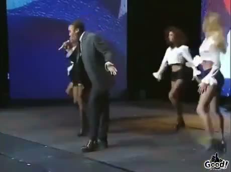 A performer on stage with a microphone gestures toward backup…