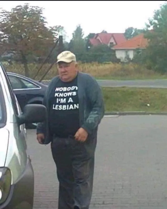 Modern Britain in a nutshell. Person wearing a cap and shirt with text standing by a car in a rural setting with houses visible in background. Text: NOBODY KNOWS I'M A LESBIAN