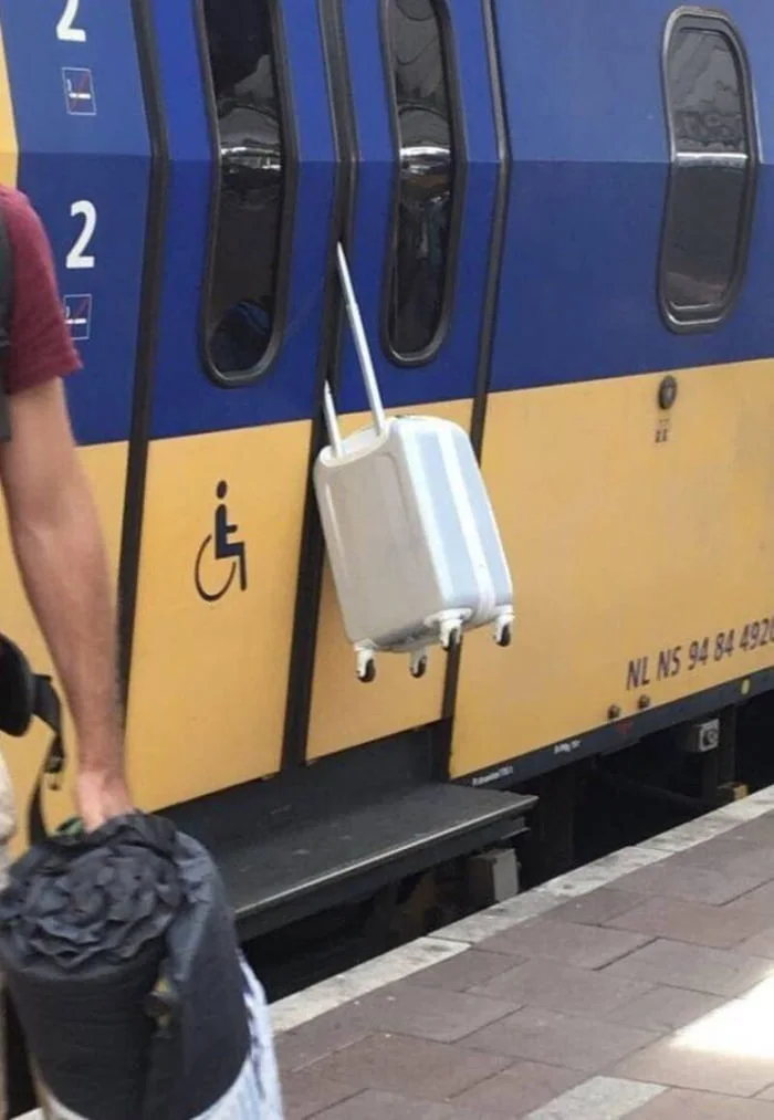Hope no tunnel till the next station. A person loading a white suitcase onto a train car with blue and yellow livery and a wheelchair accessibility symbol. Text: 2 / NL NS 94 94 4827