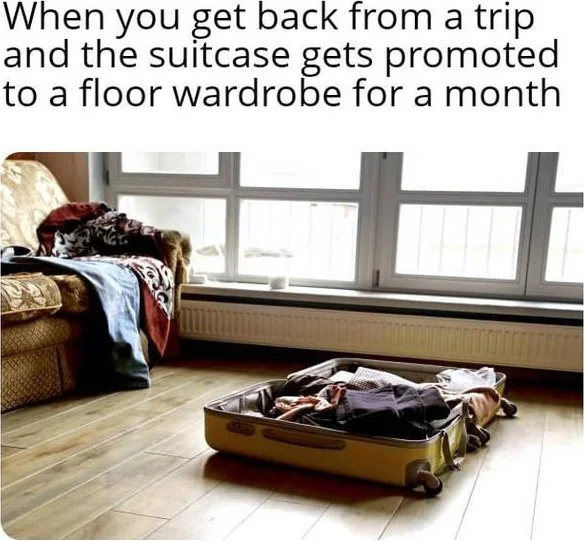 A packed suitcase sits open on a bedroom floor while clothes are piled on a nearby chair and bed. Text: When you get back from a trip and the suitcase gets promoted to a floor wardrobe for a month