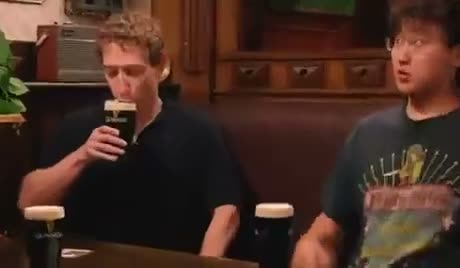 Two men sitting at a bar, one holding a microphone, appearing to be…