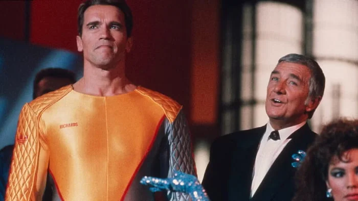 Enough with the firing squads! It’s 2026—we want the *Running Man* you promised us!. Man in orange futuristic suit stands next to man in tuxedo at formal event. Text: RICHARDS