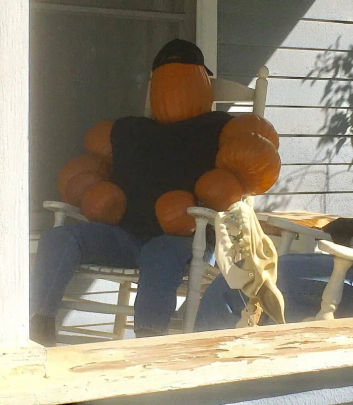 A figure made of pumpkins wearing a black shirt and cap sits in a chair on a porch