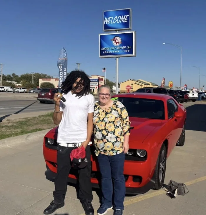 That's one way to get a Challenger. Two people posing in front of a red car at a car dealership lot with signage visible. Text: WELCOME / RIDGE CITY MOTOR LLC / (620)655-1166